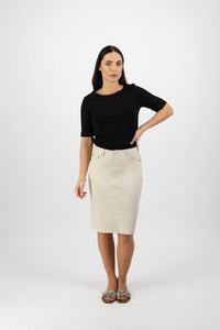 A woman with long dark hair poses against a white background, wearing a black short-sleeve top, Vassalli’s Poppy Poplin Skirt in Sand, and patterned flats. One hand rests on her hip.