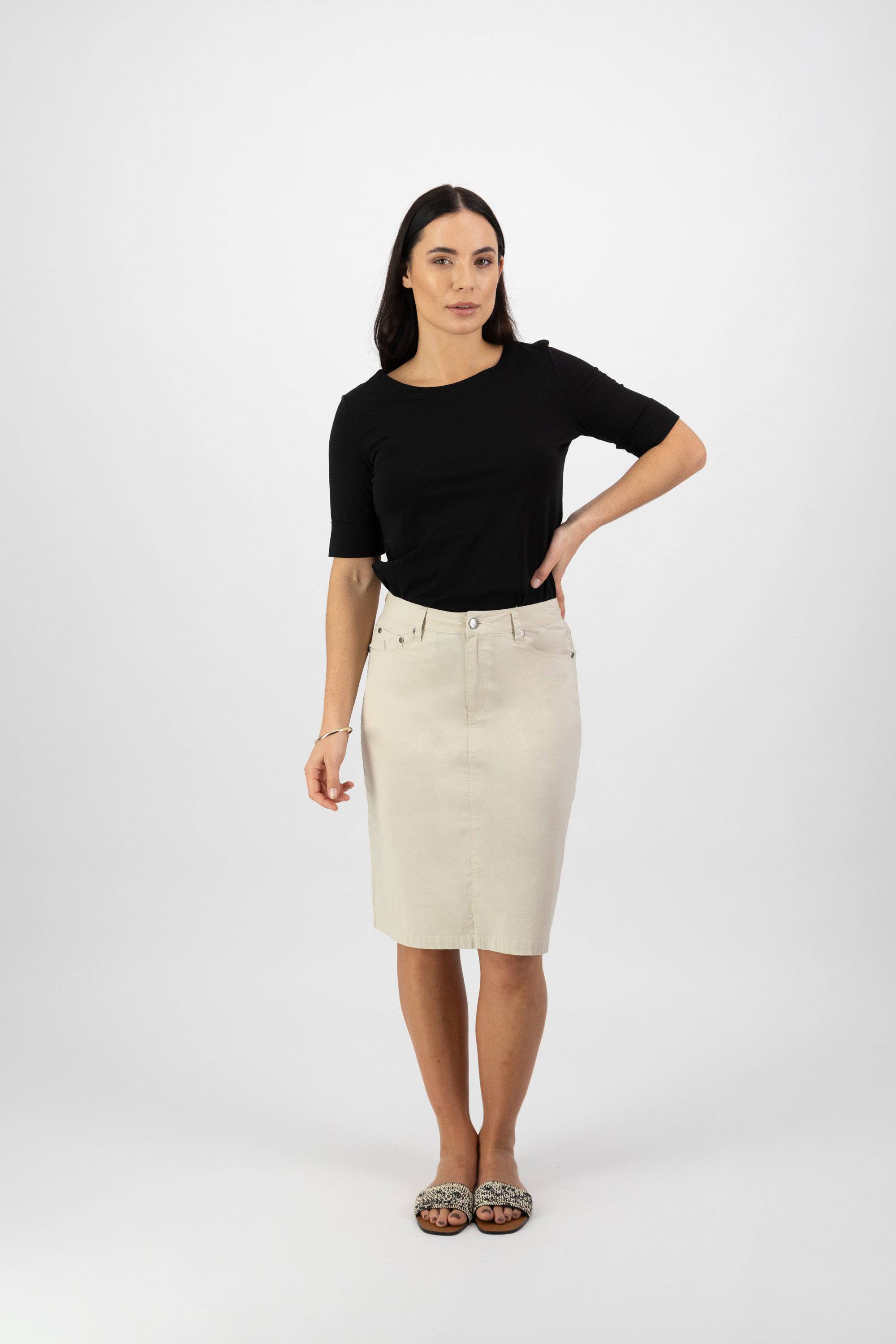 A woman with long dark hair poses against a white background, wearing a black short-sleeve top, Vassalli’s Poppy Poplin Skirt in Sand, and patterned flats. One hand rests on her hip.