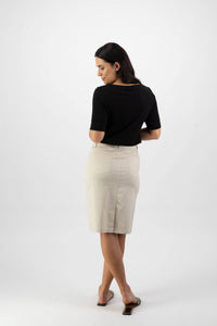 A woman faces away, looking over her shoulder, wearing a black top and the Vassalli Poppy Poplin Skirt - Sand with jean styling, plus open-toed shoes, against a white background.