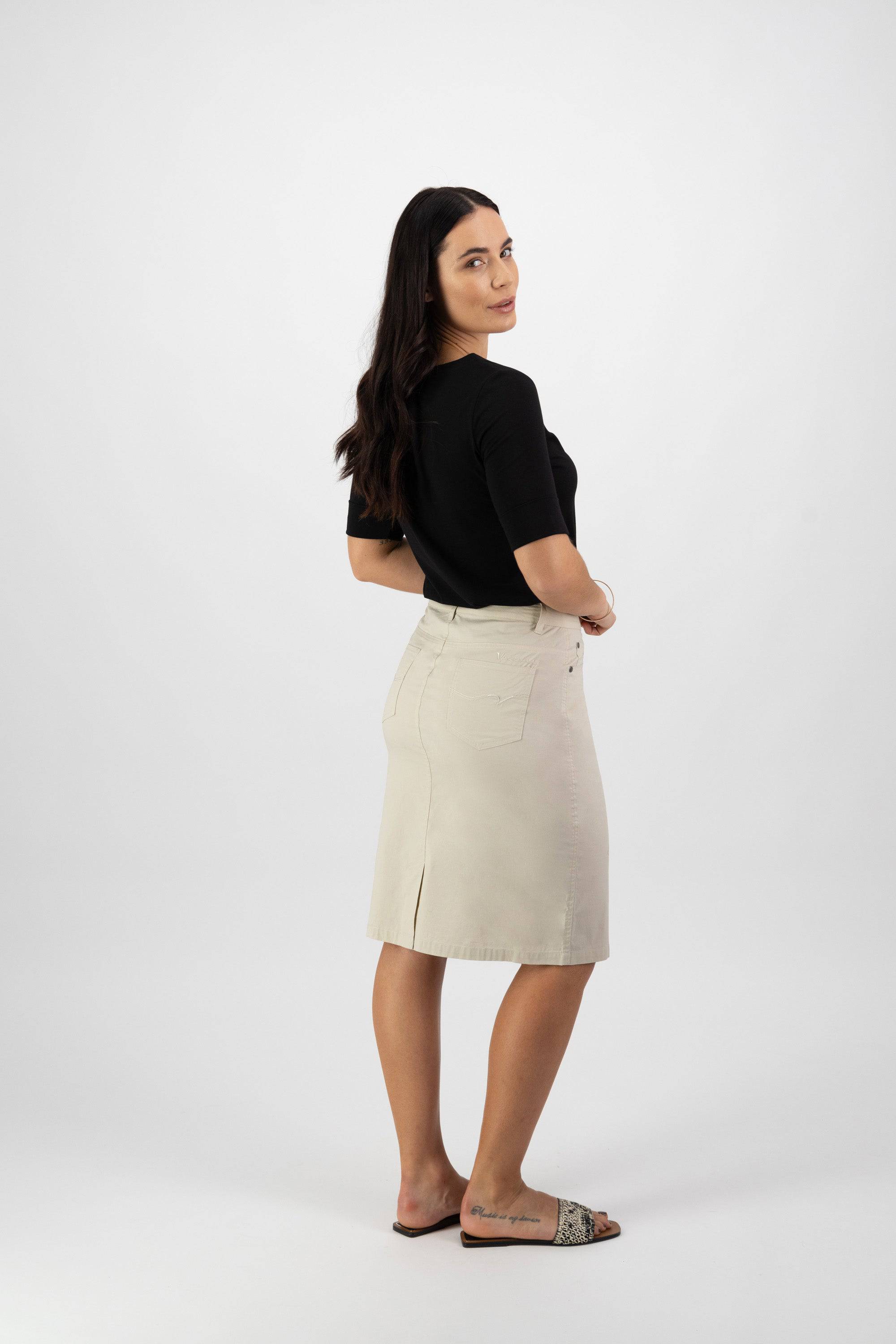 A woman with long dark hair, in a black top and sandals, models the Vassalli Poppy Poplin Skirt in Sand. She stands against a plain white background, facing slightly away and looking over her shoulder with a neutral expression.