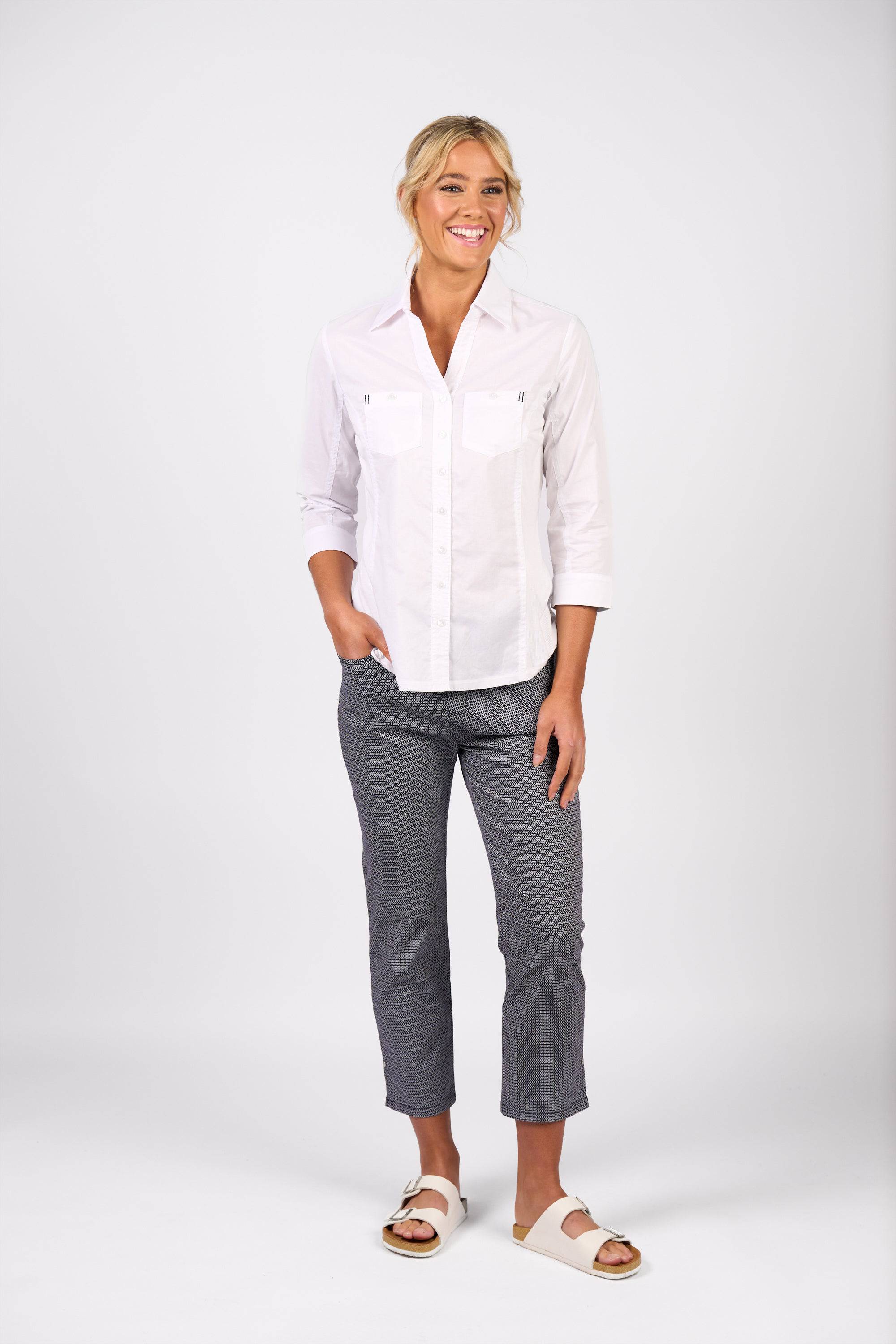 A woman smiles against a plain white background, wearing the Vassalli Isla Shirt in white. She pairs it with patterned cropped pants and white slide sandals, with one hand in her pocket and the other resting by her side.
