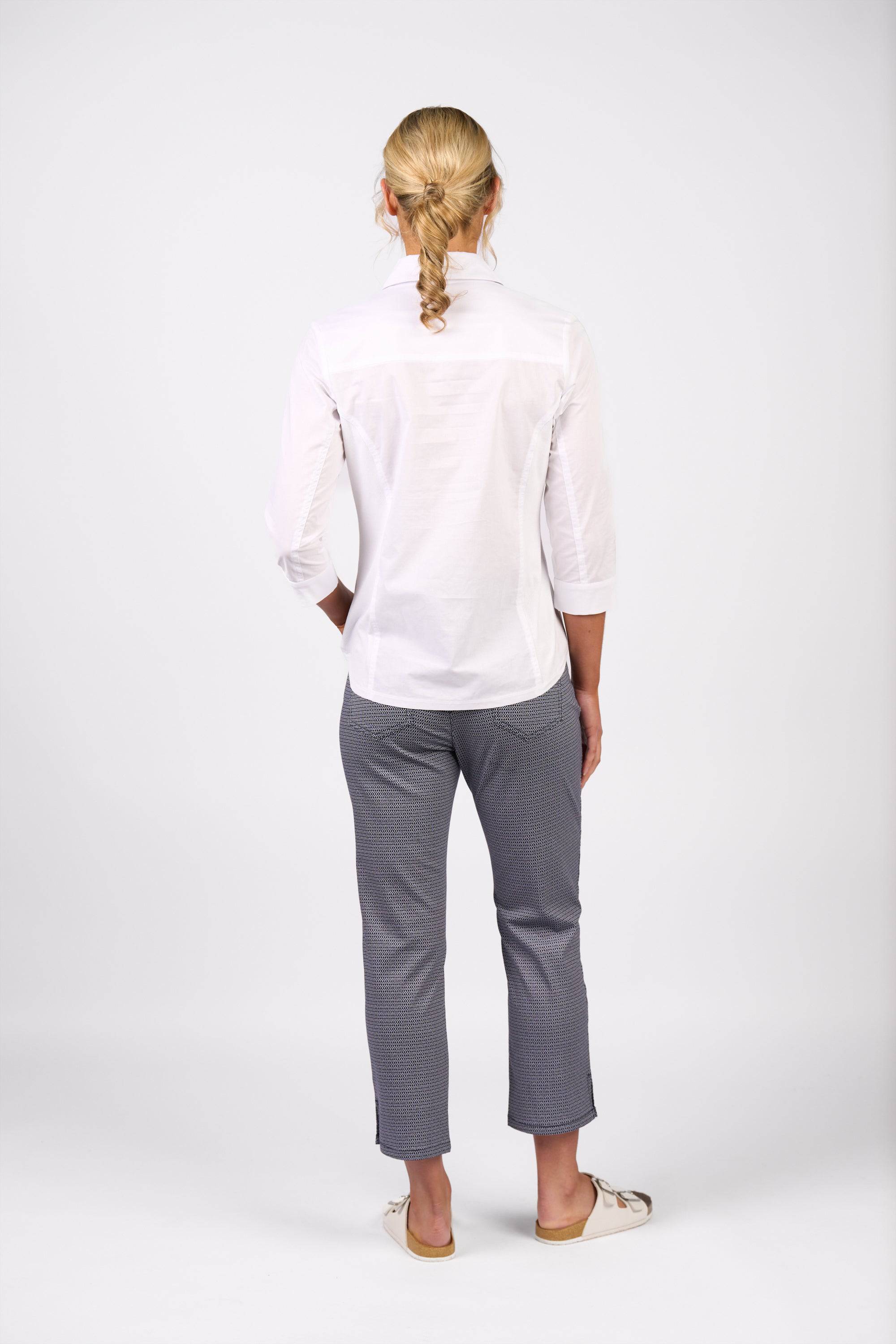 A person with braided blonde hair faces away, wearing the Vassalli Isla Shirt in white, gray pants, and beige shoes against a plain white background.