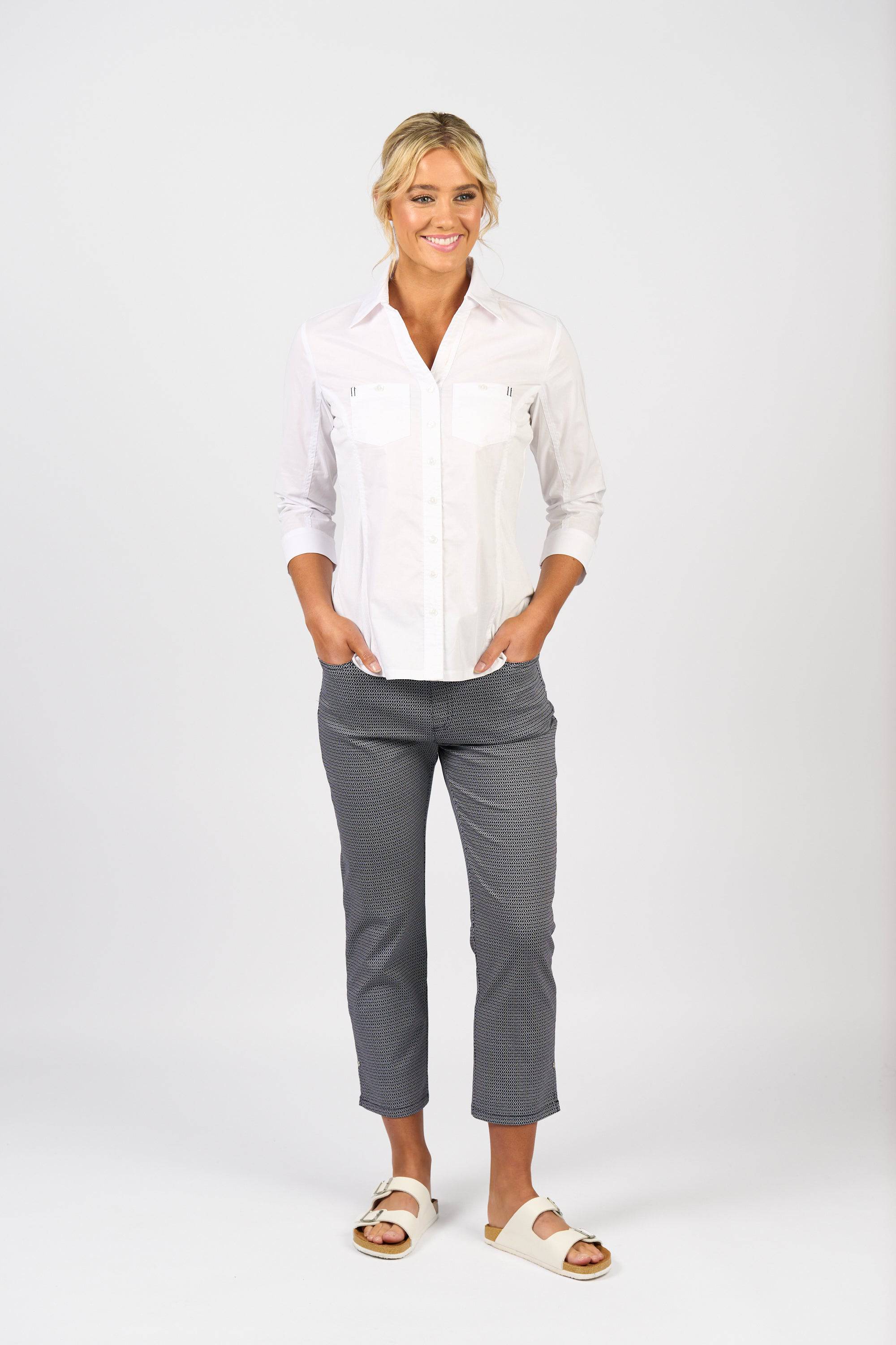 A woman smiles with hands in her pockets, wearing the Vassalli Isla Shirt in white, gray patterned pants, and white sandals against a plain white background.