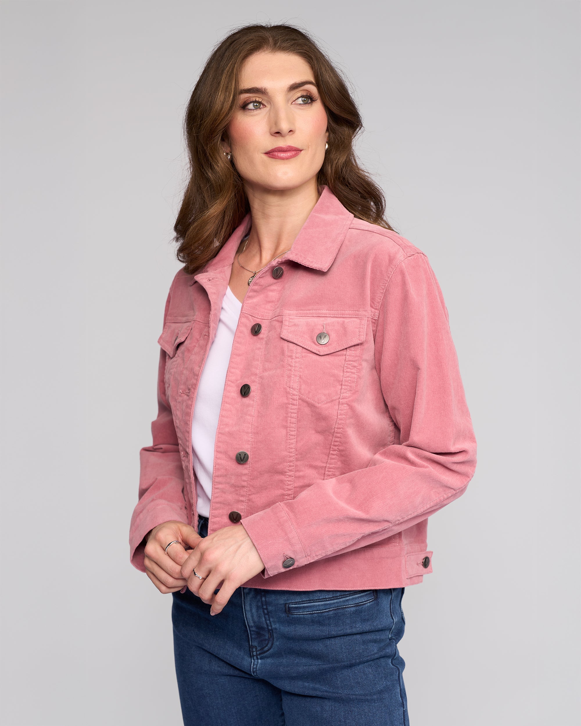 A woman with long brown hair models the Vassalli Core Cord Jacket in dusky pink over a white shirt and blue jeans, standing against a light gray background and looking slightly to her left with a slight smile.