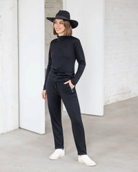 A woman smiles indoors on a concrete floor, wearing the Vassalli Riley Merino Polo in black with black pants, a black hat, and white sneakers. She stands near a white wall.
