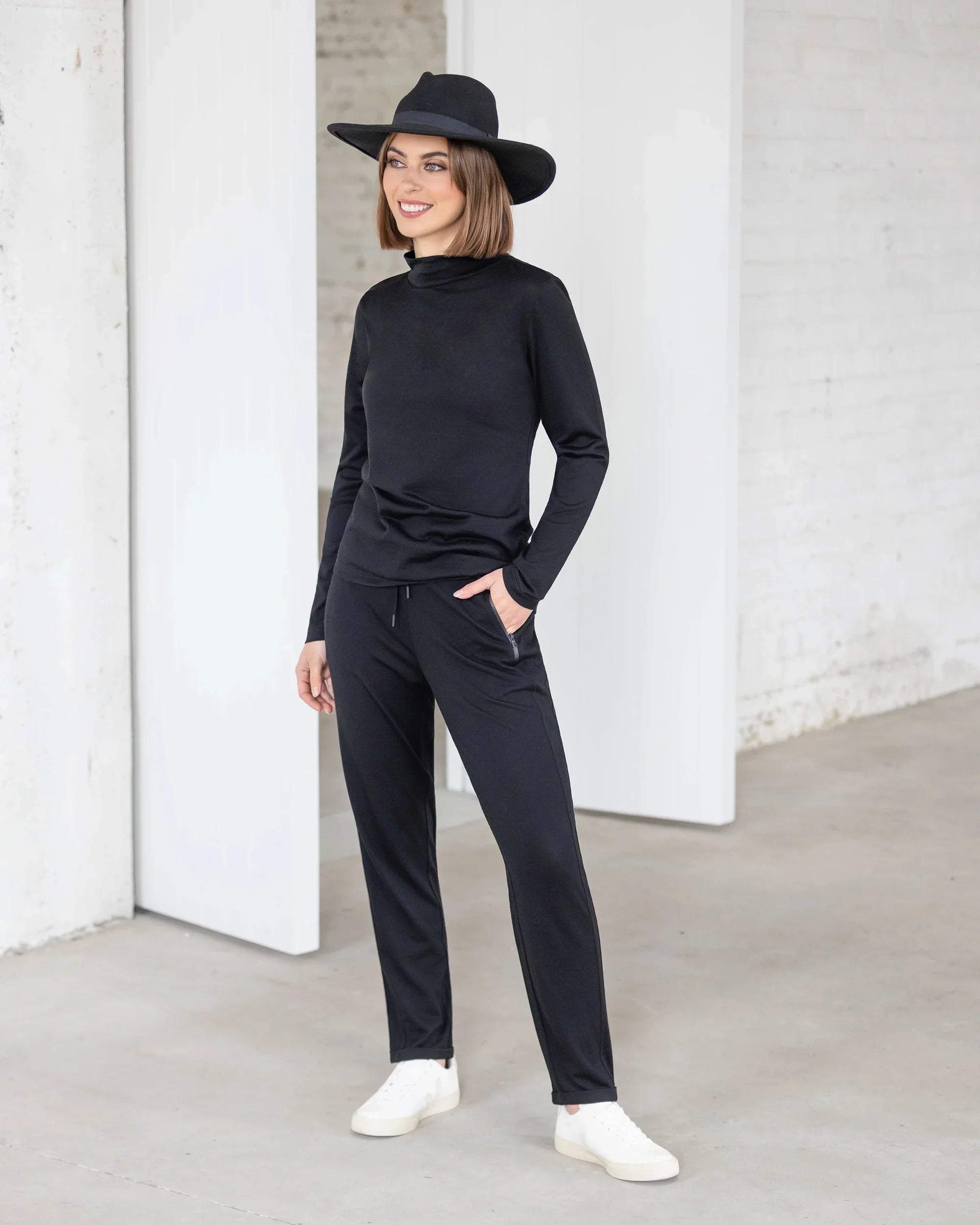 A woman smiles indoors on a concrete floor, wearing the Vassalli Riley Merino Polo in black with black pants, a black hat, and white sneakers. She stands near a white wall.