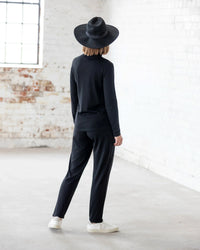 A person stands indoors, facing away, wearing the Vassalli Riley Merino Polo in black with black pants and white shoes. White brick walls and large windows highlight this versatile layering piece.