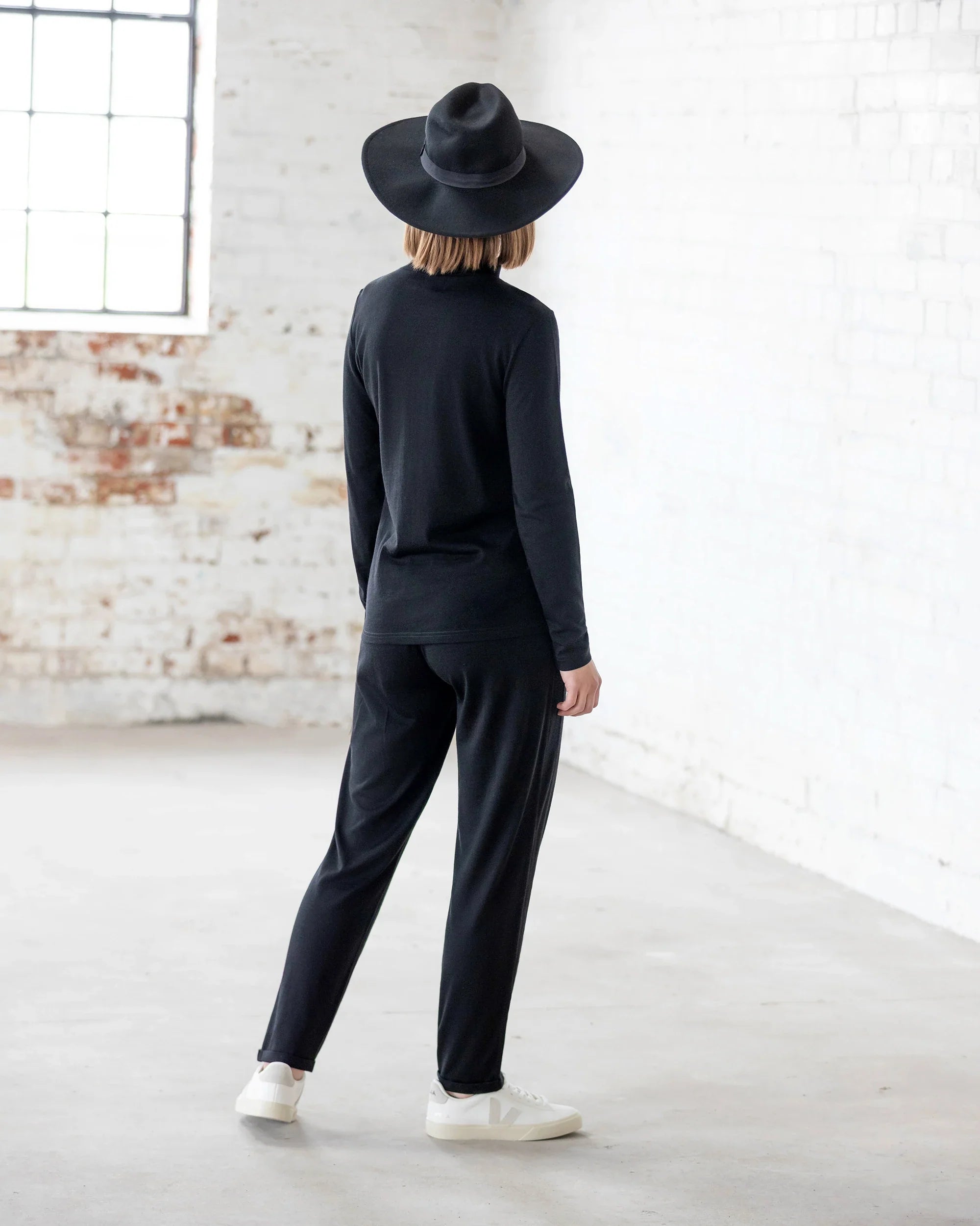 A person stands indoors, facing away, wearing the Vassalli Riley Merino Polo in black with black pants and white shoes. White brick walls and large windows highlight this versatile layering piece.