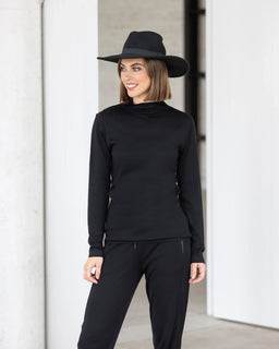 A woman wearing the Vassalli Riley Merino Polo in black and black pants stands indoors, smiling and looking to the side. White walls and columns are visible in the background.