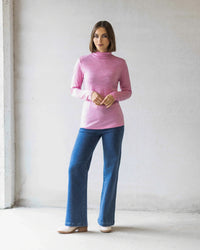 A woman stands indoors against a light wall, wearing the Vassalli Riley Merino Polo in Pink Marle with blue jeans and beige shoes. She faces forward with her hands clasped—a perfect winter layer.