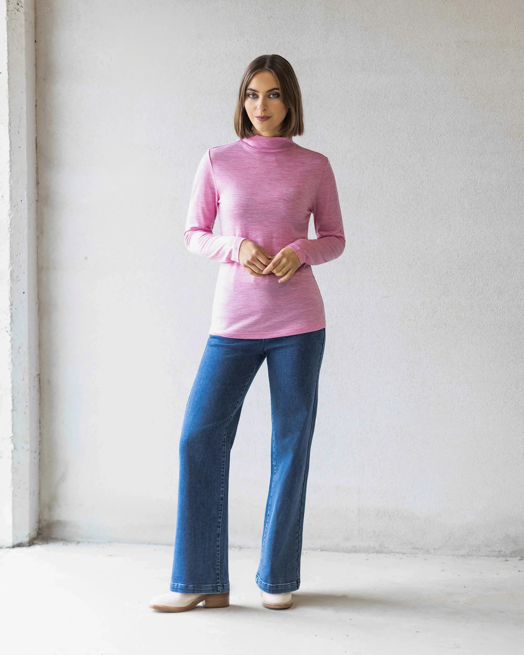 A woman stands indoors against a light wall, wearing the Vassalli Riley Merino Polo in Pink Marle with blue jeans and beige shoes. She faces forward with her hands clasped—a perfect winter layer.