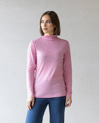 A woman with straight brown hair wears the Vassalli Riley Merino Polo in Pink Marle with blue jeans, standing against a light gray textured wall and looking slightly to her left.