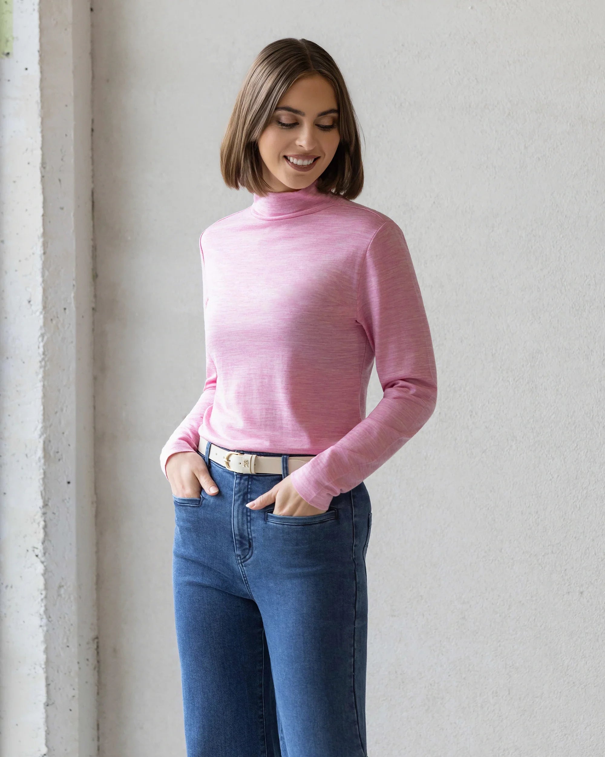 A woman with a bob haircut smiles with her hands in her pockets, wearing the Vassalli Riley Merino Polo in Pink Marle and blue jeans, standing against a light-colored wall.