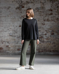 A young woman stands by a weathered brick wall, wearing the Vassalli Ivey Merino Top in black, relaxed olive green pants, and white sneakers. She gazes to the side with a casual and relaxed posture.