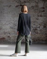 A woman with straight, shoulder-length hair faces a brick wall, wearing the Vassalli Ivey Merino Top in black, olive green pants, white sneakers, and holding a small black handbag.