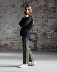A woman with straight brown hair stands sideways by a brick wall, wearing the Vassalli Ivey Merino Top in black, green pants, white sneakers, and carrying a black shoulder bag.