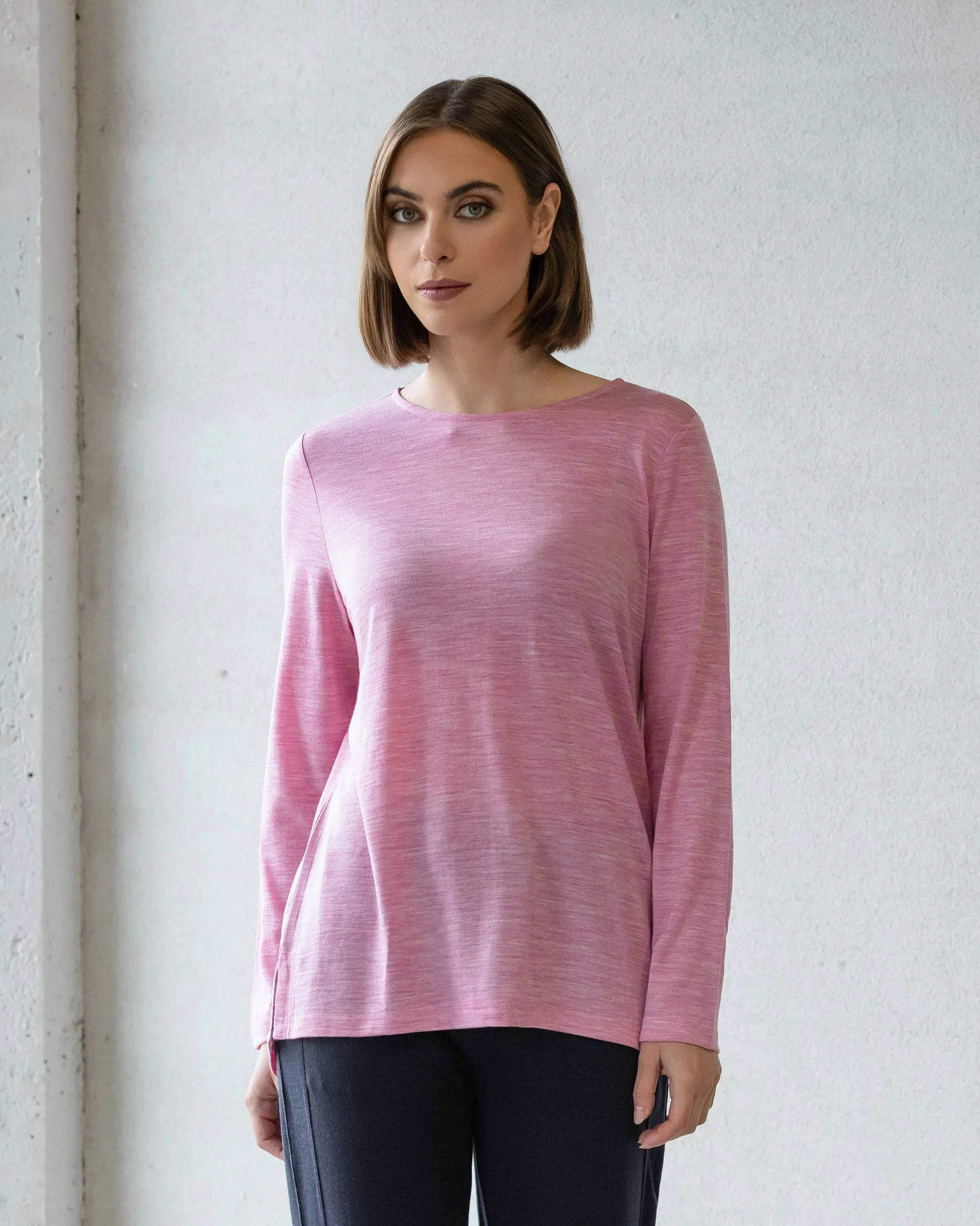 A woman with straight, shoulder-length brown hair wears the Vassalli Ivey Merino Top in Pink Marle and dark pants, standing against a light wall and looking at the camera—an ideal winter layering piece.