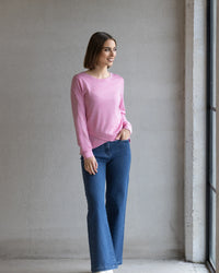 A woman with a bob stands indoors near a window, smiling. She’s wearing the Vassalli Remy Merino Top in Pink Marle and blue jeans—ideal for winter layering—with one hand in her pocket against a plain gray wall.