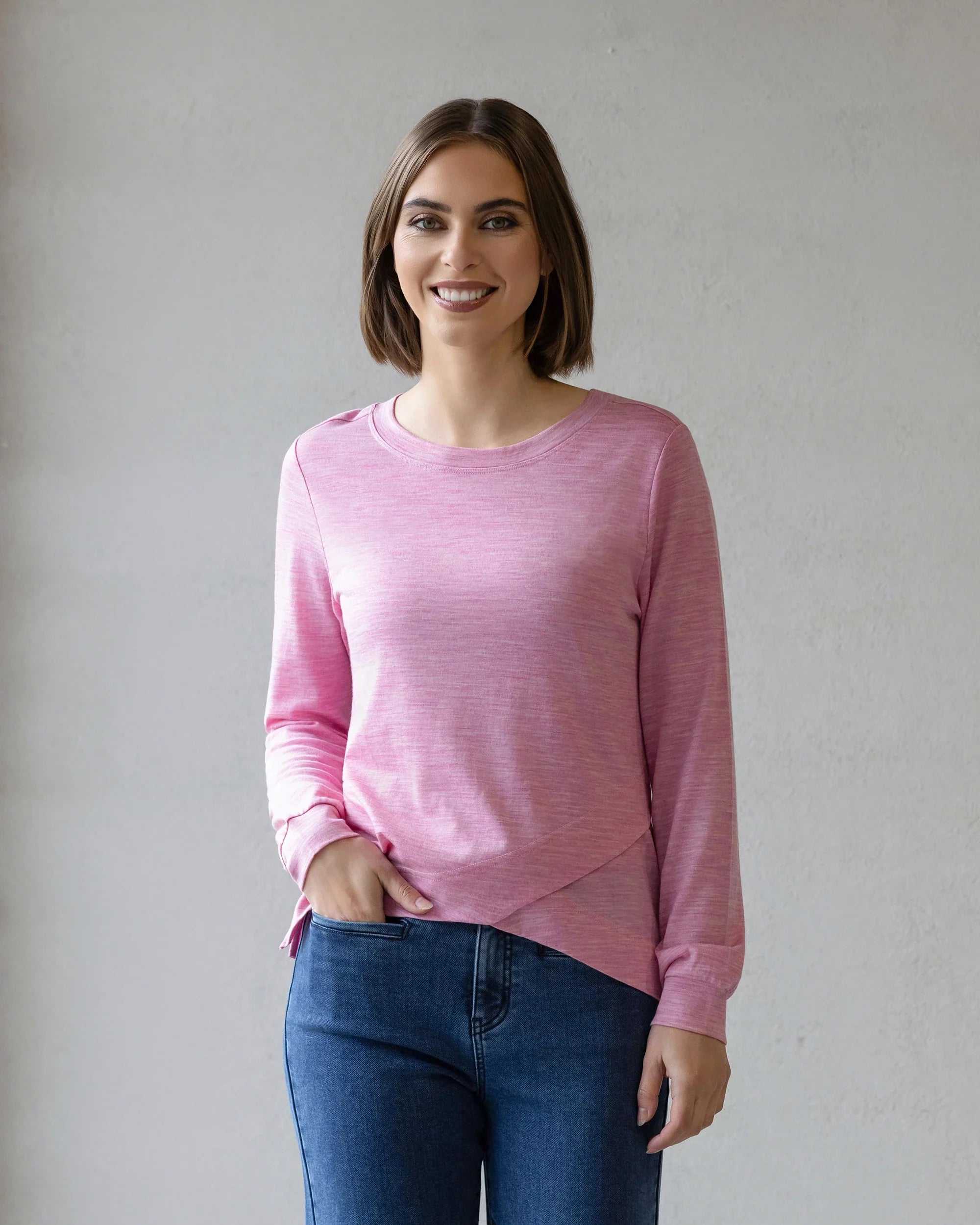 A woman with short brown hair smiles at the camera, wearing the Vassalli Remy Merino Top in Pink Marle and blue jeans, ideal for winter layering, while standing against a plain light gray background.