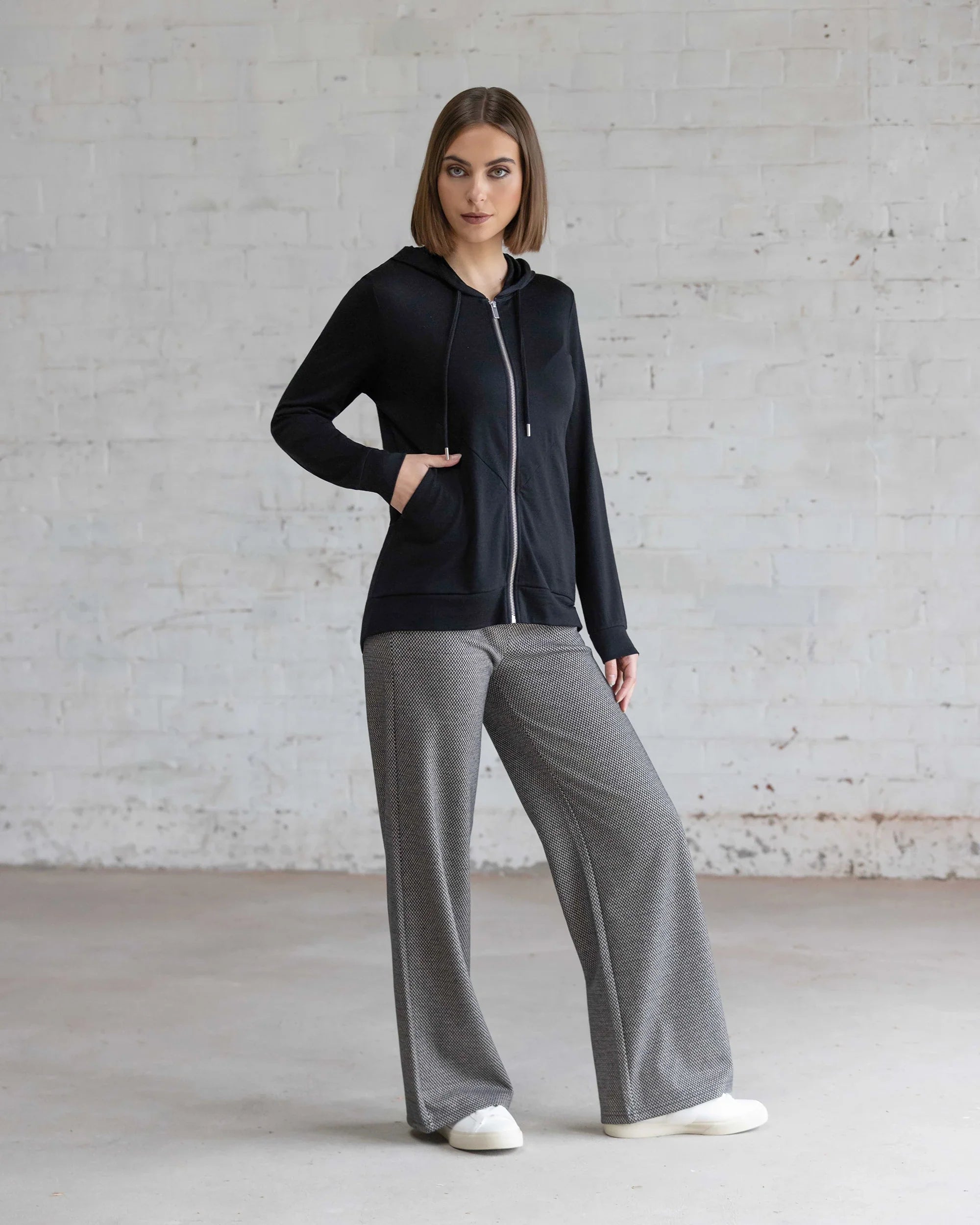 A woman stands indoors against a white brick wall, wearing the Vassalli Shield Merino Hoodie in black, paired with gray wide-leg pants and white shoes. One hand is in her pocket, the other rests on her thigh.