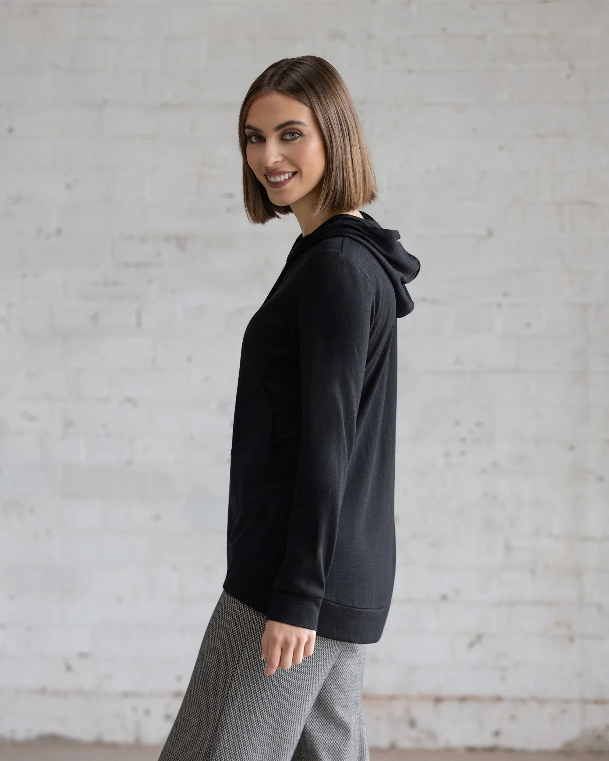 A woman with a bob haircut poses sideways, smiling in front of a white brick wall, wearing the Vassalli Shield Merino Hoodie in black and gray checkered pants.