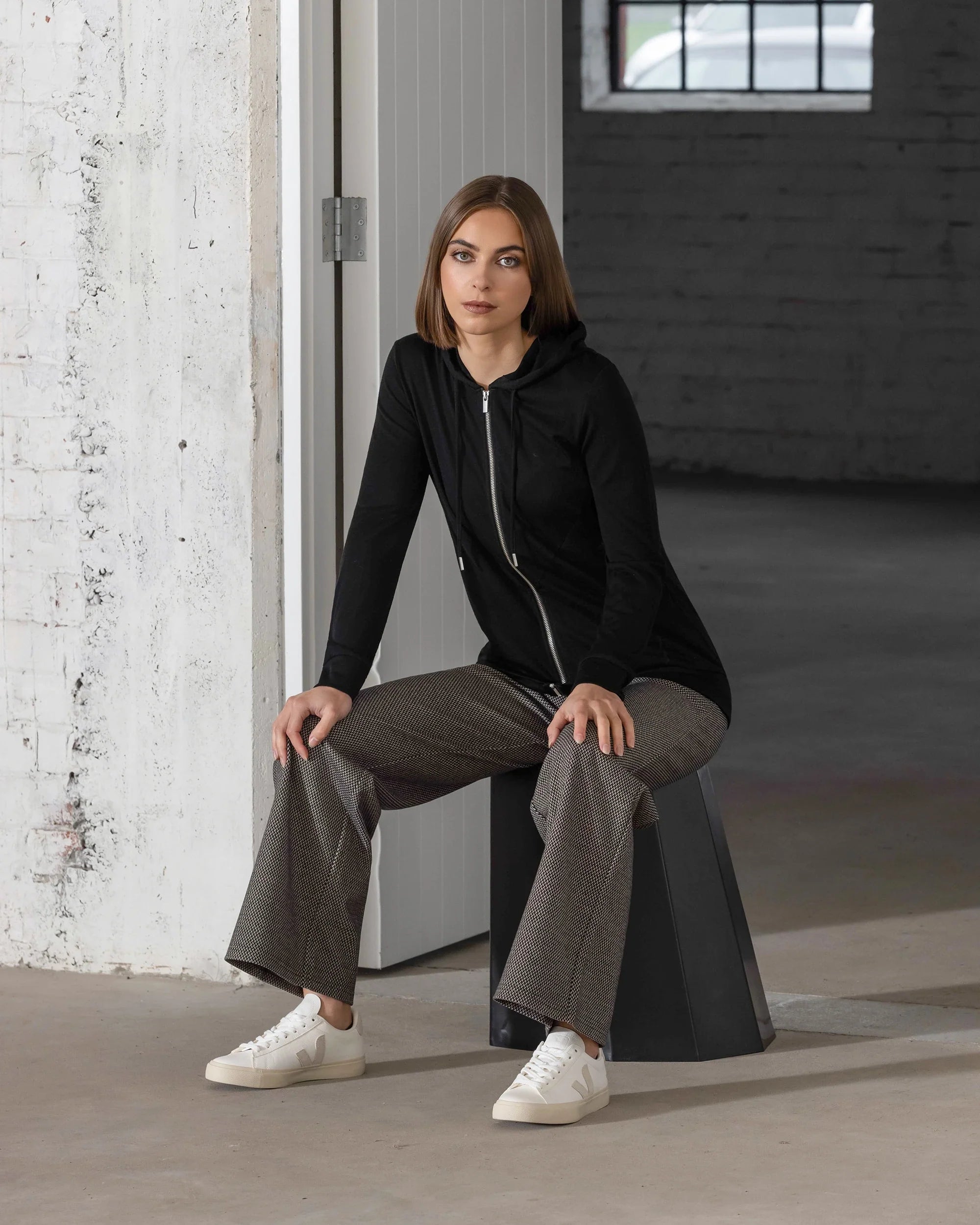 A woman with straight, shoulder-length brown hair sits on a dark angular stool in a minimalist industrial space, wearing Vassalli's Shield Merino Hoodie in black, gray plaid wide-leg pants, and white sneakers as she looks calmly at the camera.