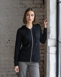 A woman with straight brown hair wears the Vassalli Shield Merino Hoodie in black and gray patterned pants, standing by a window with one hand on a concrete wall, her sleek hoodie contrasting against the white brick wall behind her.