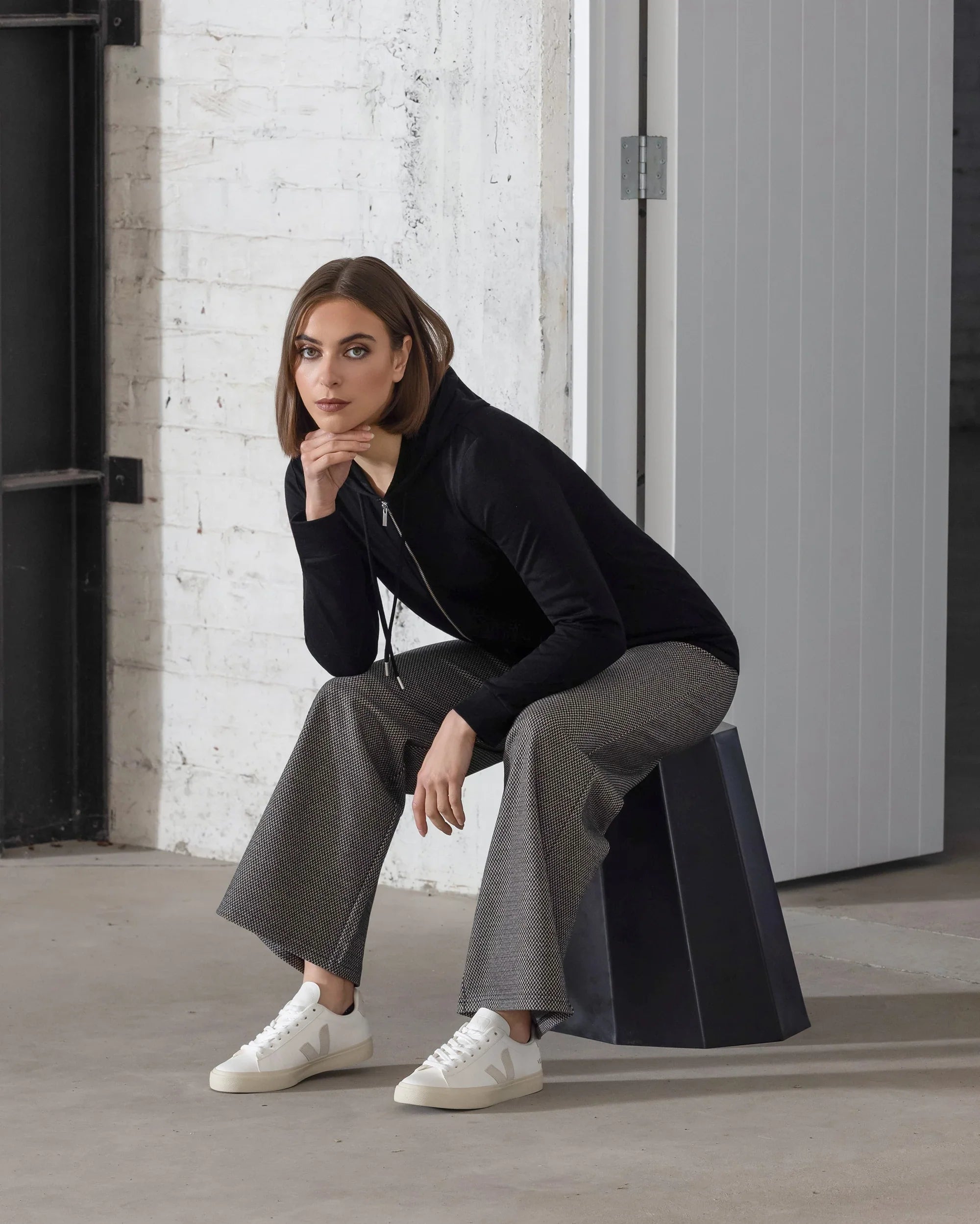 A woman with straight brown hair wears the Vassalli Shield Merino Hoodie in black, styled with gray wide-leg pants and white sneakers, seated on a modern black stool in a minimalist industrial room with white brick walls.