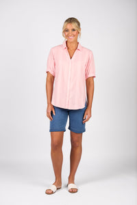 A woman stands against a plain white background, smiling. She wears a Vassalli Shirt - Pink Stripe, denim knee-length shorts, and white slide sandals.