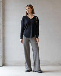 A woman stands indoors against a light-colored wall, smiling. She wears the Vassalli Veni Merino Top in black, gray wide-leg pants, black shoes, and carries a shoulder bag.
