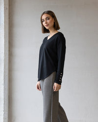 A woman with straight, shoulder-length brown hair stands indoors against a light gray wall, wearing the Vassalli Veni Merino Top in black and gray patterned pants. She faces the camera with a slight smile.