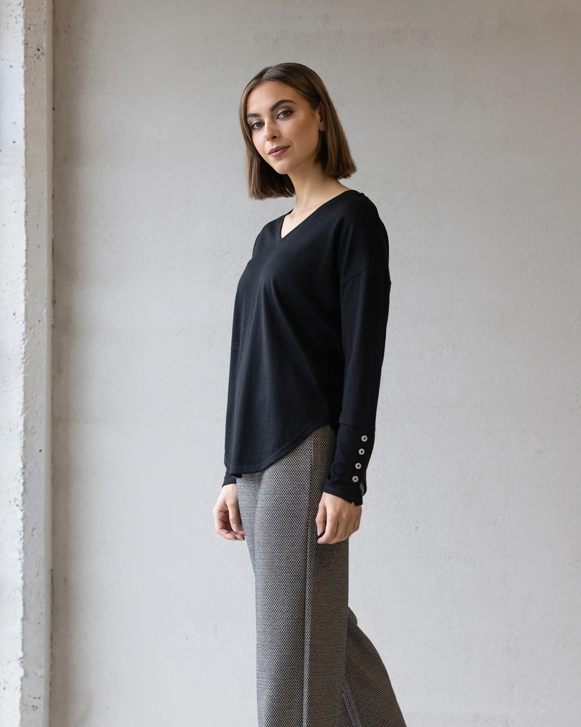 A woman with straight, shoulder-length brown hair stands indoors against a light gray wall, wearing the Vassalli Veni Merino Top in black and gray patterned pants. She faces the camera with a slight smile.
