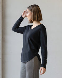 A woman with a straight brown bob stands sideways, looking left with her right hand near her forehead, wearing the Vassalli Veni Merino Top in black and gray patterned pants against a plain light background.