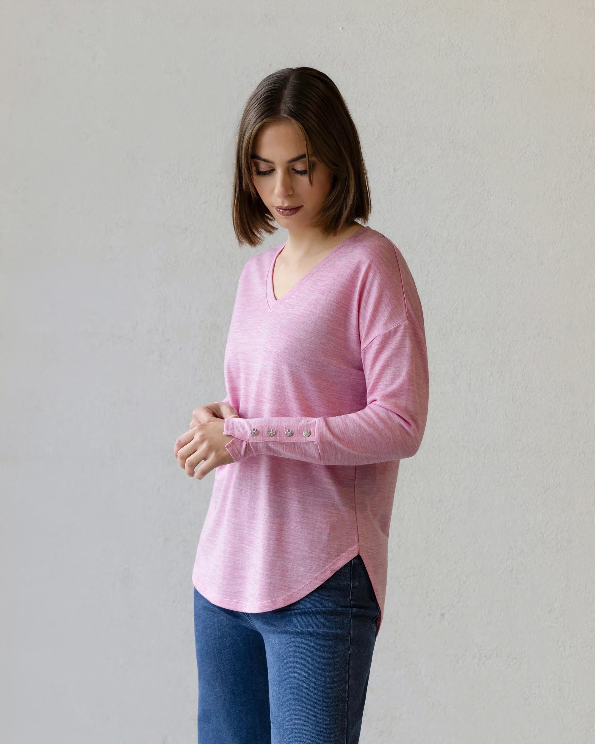 A woman with shoulder-length brown hair wears the Vassalli Veni Merino Top in Pink Marle and blue jeans, standing against a plain light background while looking down and touching her hands—perfect for winter layering.