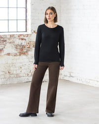 A woman with short brown hair stands indoors near a white brick wall and window, wearing the Vassalli Tori Merino Crew in black, paired with brown wide-leg pants and black shoes. She has a neutral expression. Ideal for effortless layering.