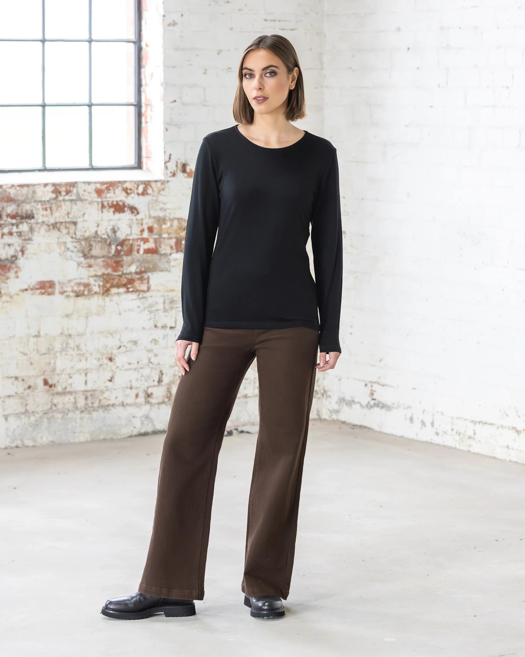 A woman with short brown hair stands indoors near a white brick wall and window, wearing the Vassalli Tori Merino Crew in black, paired with brown wide-leg pants and black shoes. She has a neutral expression. Ideal for effortless layering.