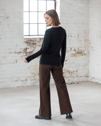 A person with short hair wears the Vassalli Tori Merino Crew in black, paired with brown wide-leg pants and black shoes, standing in a bright industrial room with white brick walls and large windows—perfect for effortless layering.