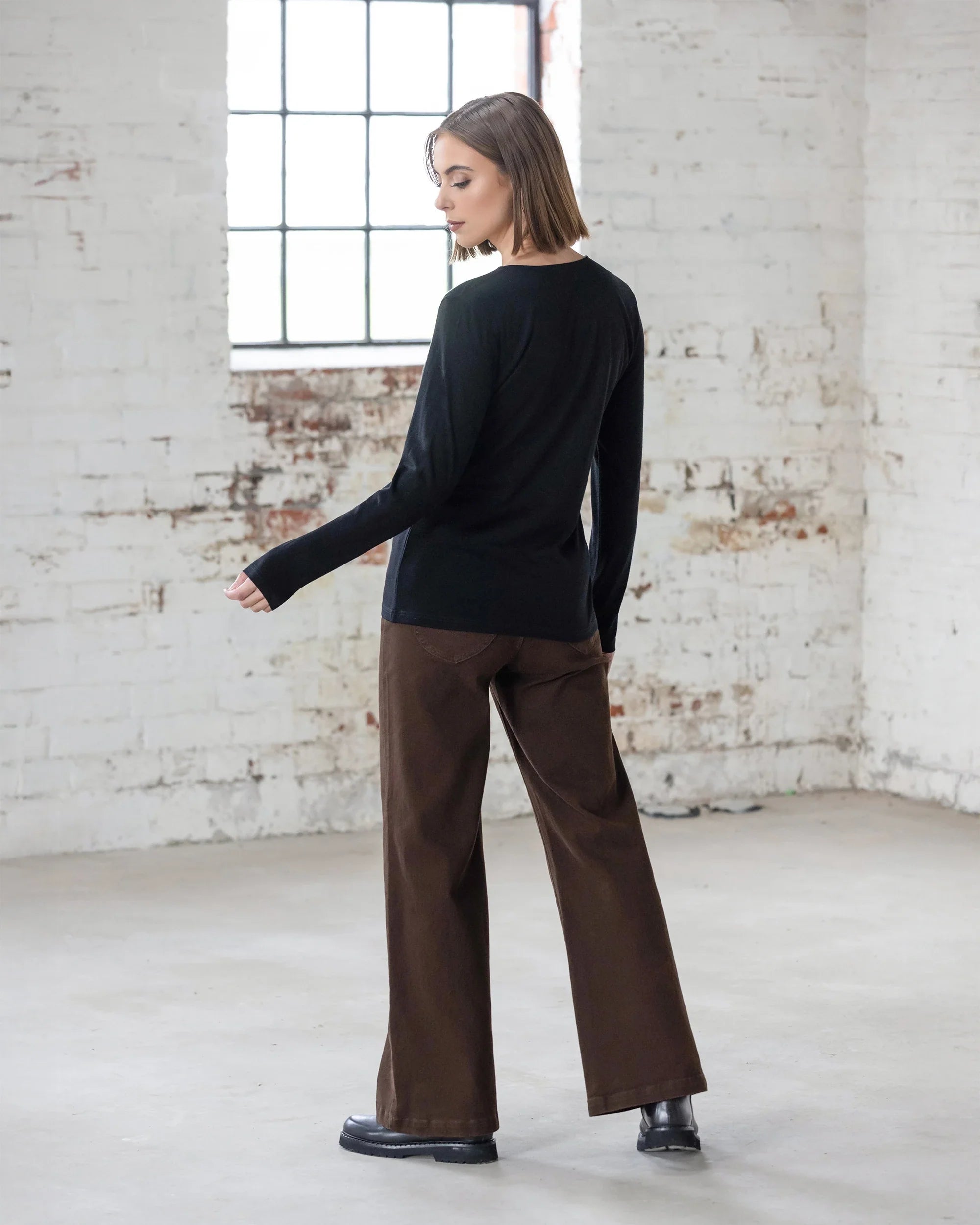A person with short hair wears the Vassalli Tori Merino Crew in black, paired with brown wide-leg pants and black shoes, standing in a bright industrial room with white brick walls and large windows—perfect for effortless layering.
