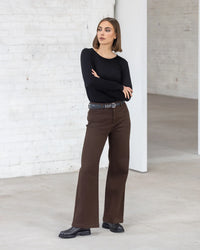 A woman stands indoors with arms crossed, wearing the Vassalli Tori Merino Crew in black, paired with brown high-waisted pants, a black belt, and black shoes. She looks to the side in a bright minimalist space with white brick walls.