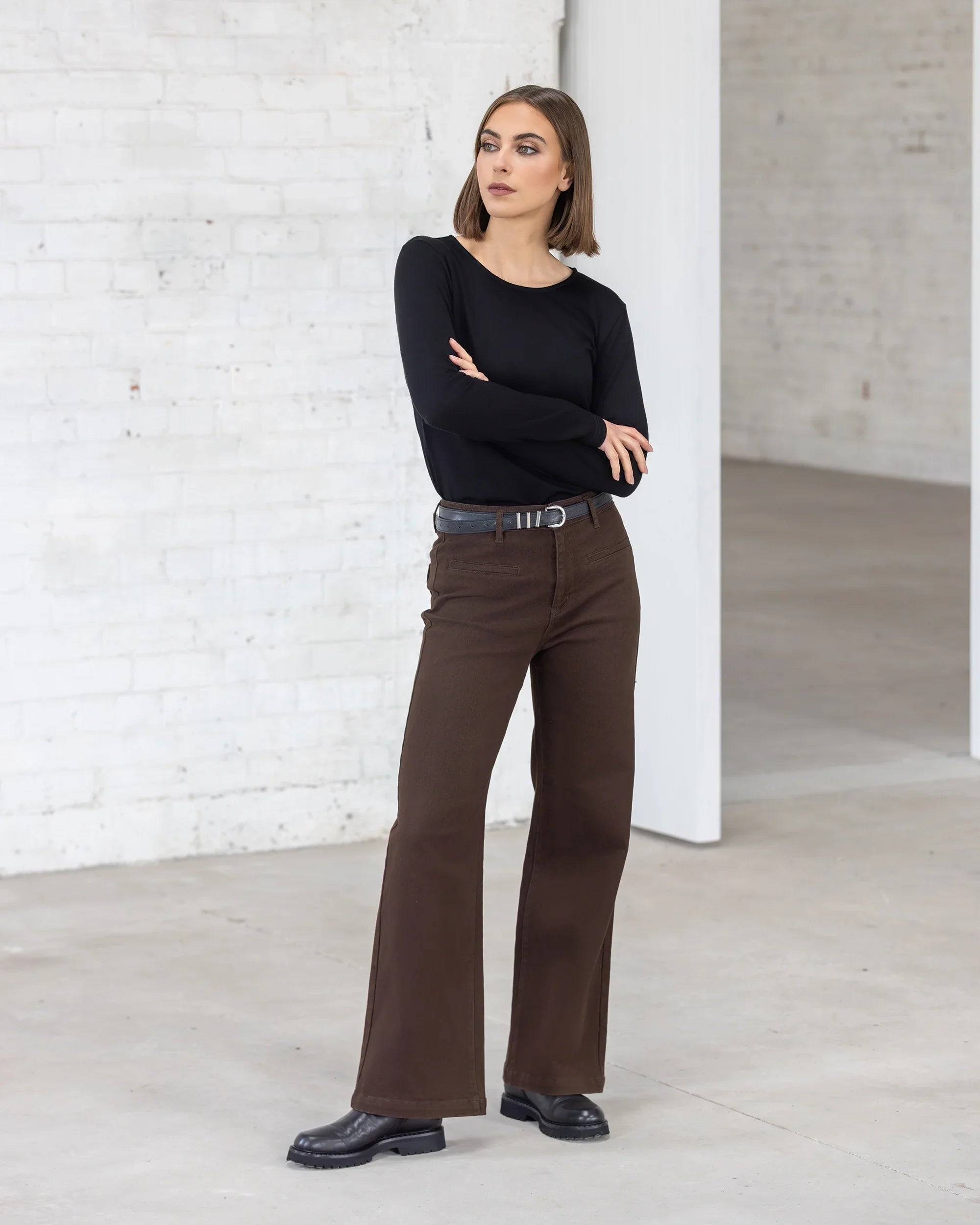 A woman stands indoors with arms crossed, wearing the Vassalli Tori Merino Crew in black, paired with brown high-waisted pants, a black belt, and black shoes. She looks to the side in a bright minimalist space with white brick walls.