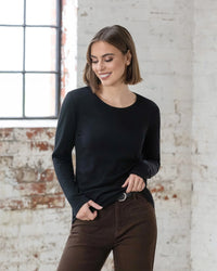 A woman with short brown hair, wearing the Vassalli Tori Merino Crew in black and brown pants, stands smiling in front of a white brick wall with a window, looking down to her left.