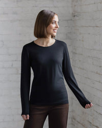 A woman with straight, shoulder-length brown hair wears the Vassalli Tori Merino Crew in black and brown pants, standing against a white brick wall and smiling slightly. Ideal for layering all year round.