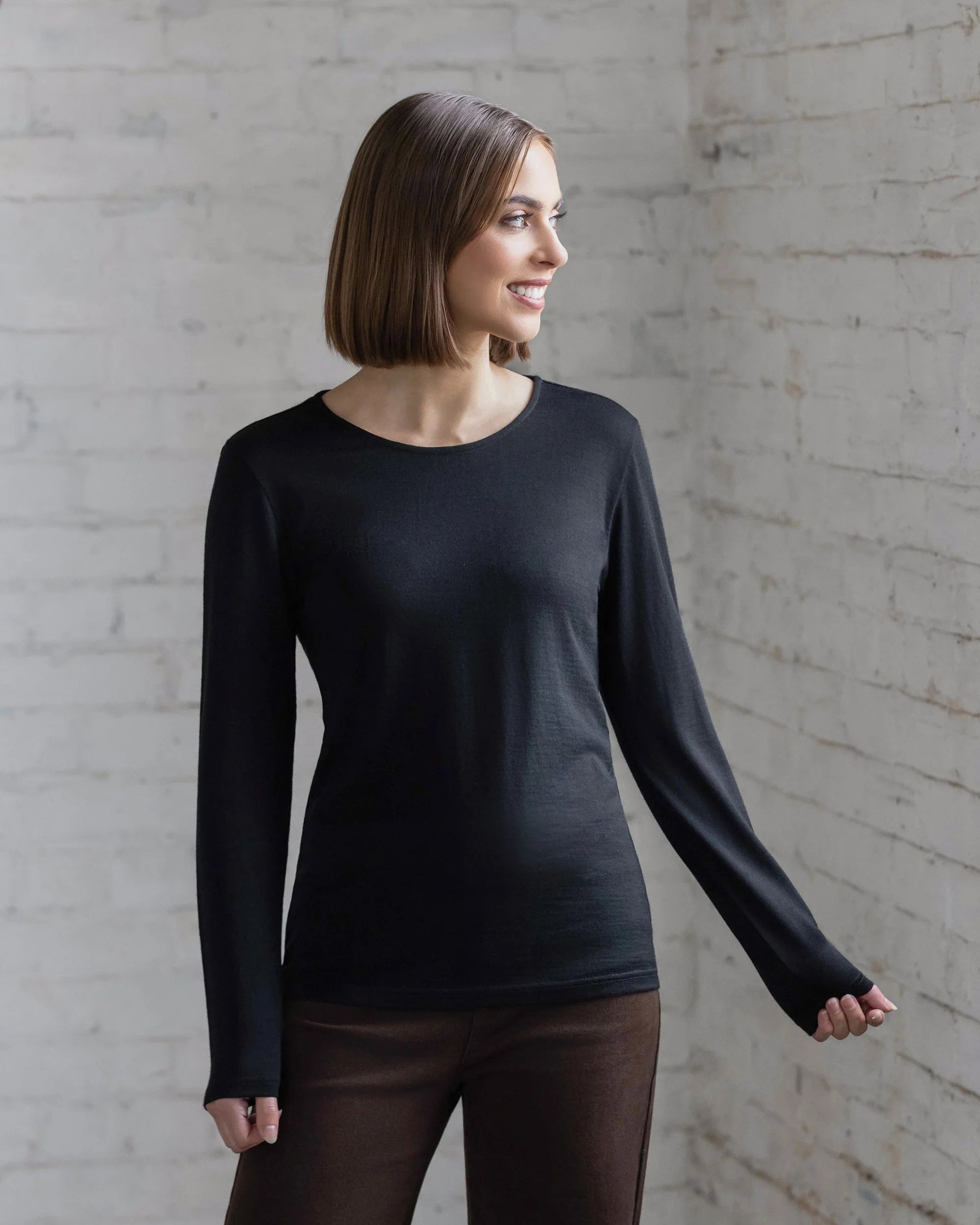 A woman with straight, shoulder-length brown hair wears the Vassalli Tori Merino Crew in black and brown pants, standing against a white brick wall and smiling slightly. Ideal for layering all year round.