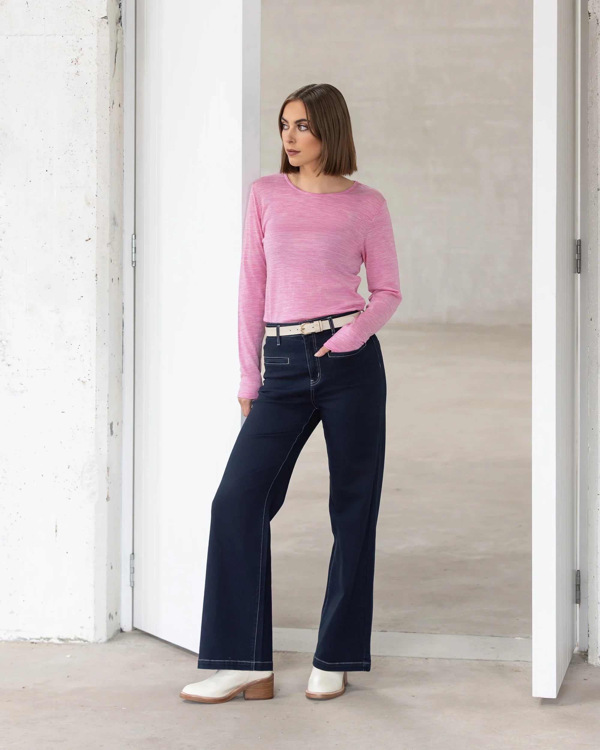 A woman with a bob haircut wears the Vassalli Tori Merino Crew in Pink Marle, paired with dark wide-leg jeans, a beige belt, and white boots. She stands casually in a minimalist white doorway—perfect for winter layering.
