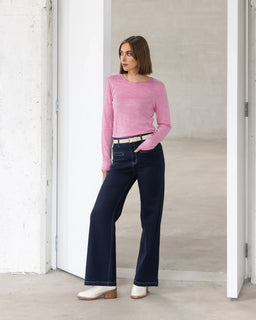 A woman with a bob haircut wears the Vassalli Tori Merino Crew in Pink Marle, paired with dark wide-leg jeans, a beige belt, and white boots. She stands casually in a minimalist white doorway—perfect for winter layering.