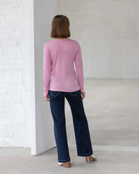 A person with straight, shoulder-length brown hair stands indoors wearing the Vassalli Tori Merino Crew in Pink Marle, paired with dark blue jeans and light shoes. The minimal white-walled setting makes it ideal for winter layering.