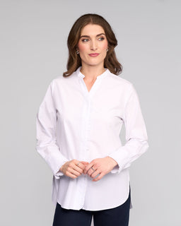 A woman with long brown hair wears the Vassalli Manhattan Shirt in white with dark pants, standing against a plain light gray background. She looks slightly left with a neutral expression and her hands clasped in front of her.