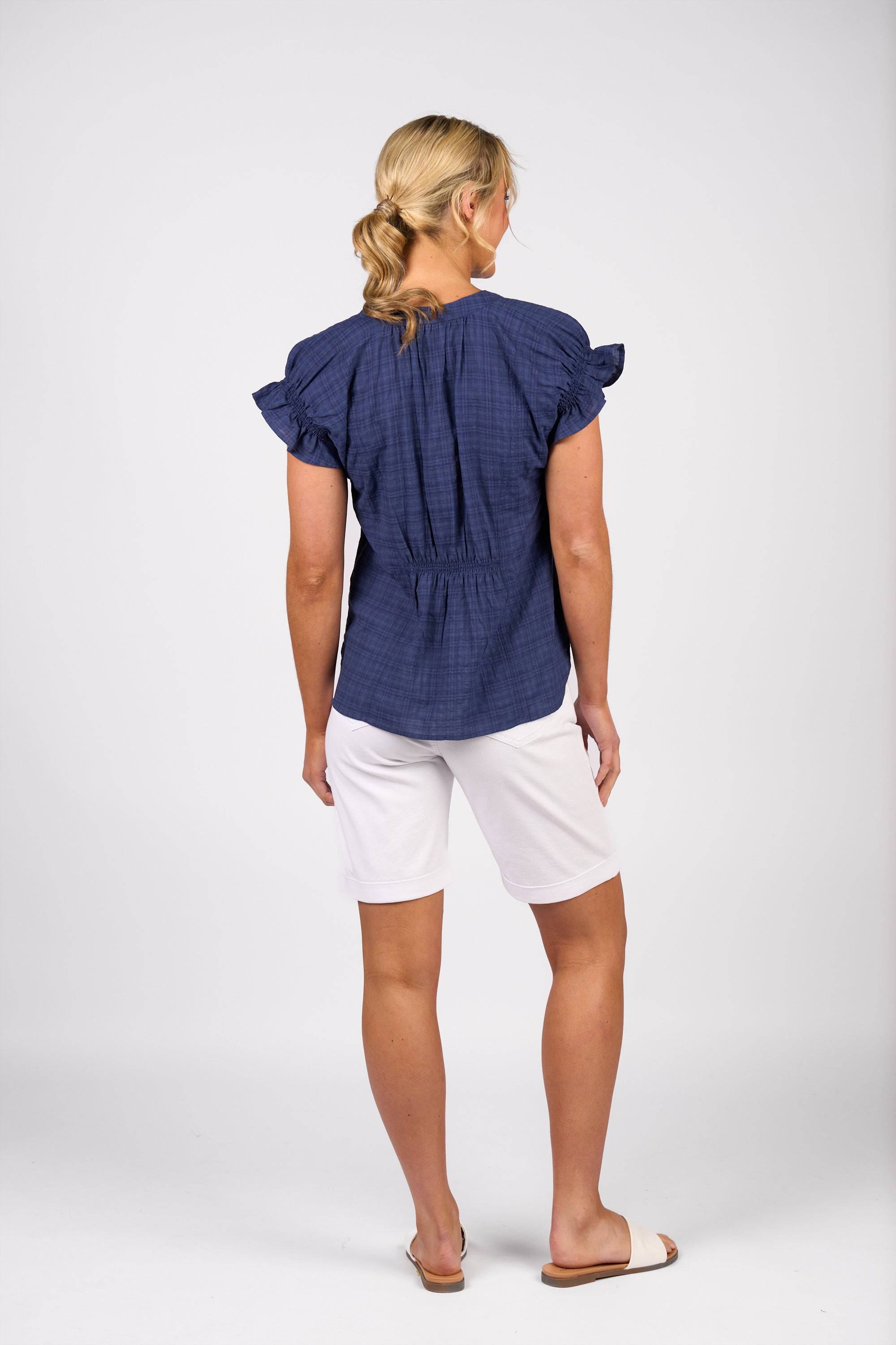 A woman with blonde hair tied back faces away, wearing the Vassalli Evelyn Top in navy with ruffled sleeves, white knee-length shorts, and white slide sandals against a plain light background.