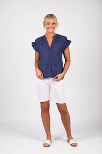 A woman stands smiling against a plain light background, wearing the Vassalli Evelyn Top in navy, white knee-length shorts, and white slide sandals. Her hair is pulled back and she poses with one hand in her pocket.