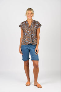 A woman stands smiling against a plain white background, wearing the Vassalli Evelyn Top - Nala with a brown and black leopard print, blue knee-length denim shorts, and tan sandals.